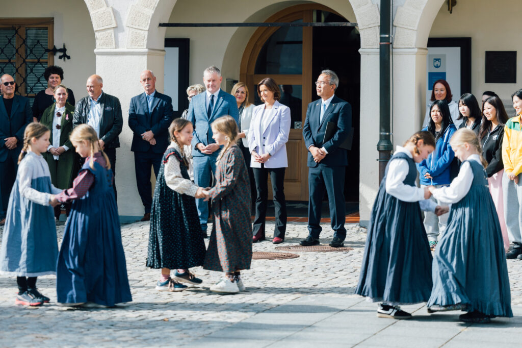 Skupina ljudi pleše na trgu, ministrica Tanja Fajon stoji in jih gleda s še nekaj drugimi ljudmi ob sebi.