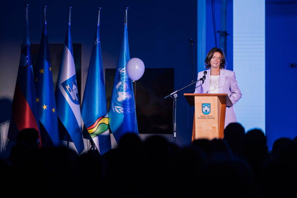 
Ministrica Tanja Fajon na desni strani stoji za govornico in nagovarja prisotne na dogodku. Na levi strani postavljene zastave.