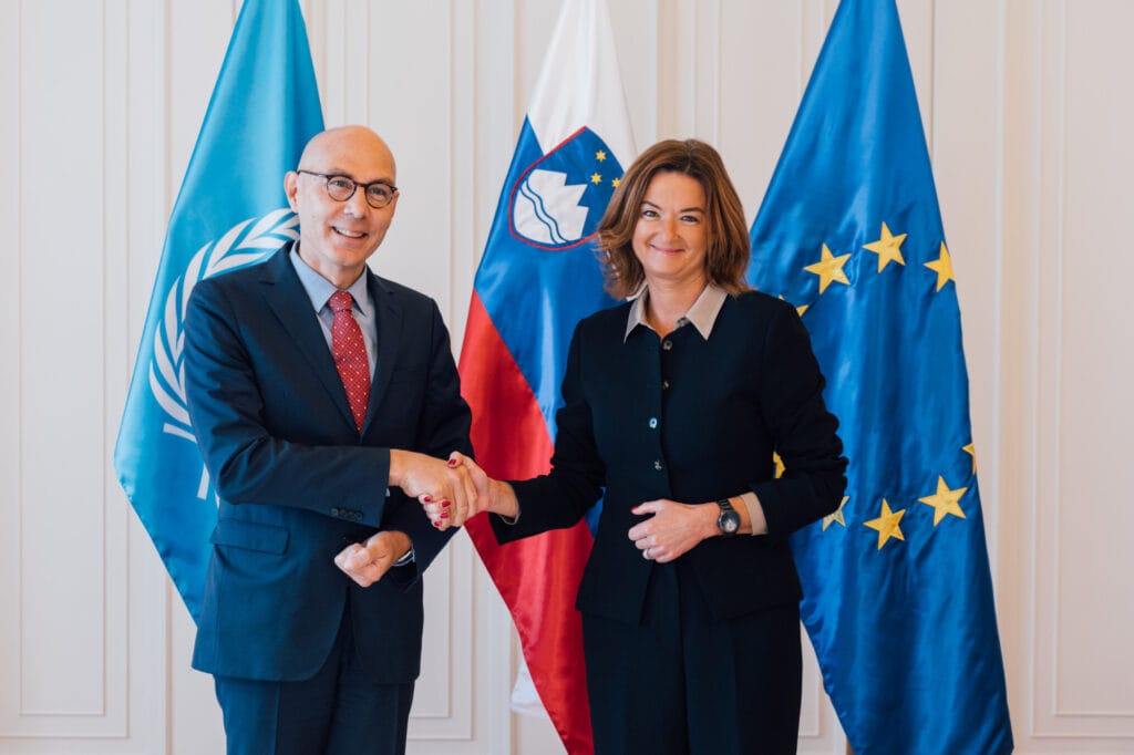 Visoki komisar OZN za človekove pravice Volker Türk in ministrica Tanja Fajon stojita pred zastavami in se rokujeta. 